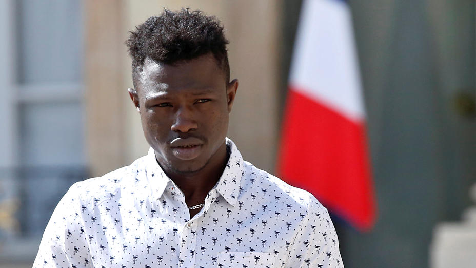 a dark-skinned man narrows his eyes, he wears a white shirt covered in black flamingoes