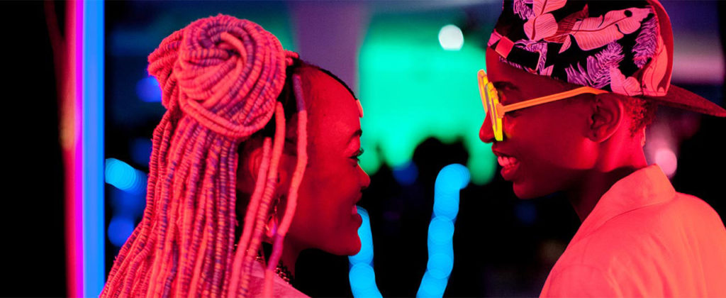 hot pink light shines over two women smiling at each other. one has long pink braids, the other is wearing a cap and sunglasses