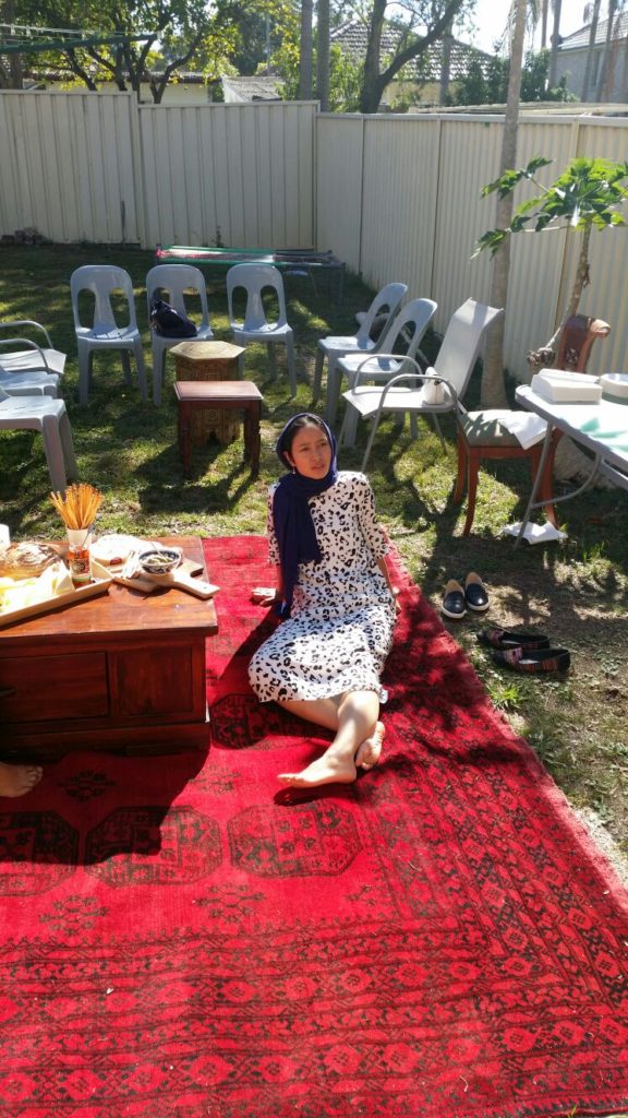 Aisyah Shah Idil with bare feet and a headscarf, leaning back on a rug in a backyard
