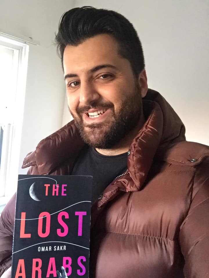 Omar Sakr with slicked back hair, wearing a down jacket, holding his book The Lost Arabs