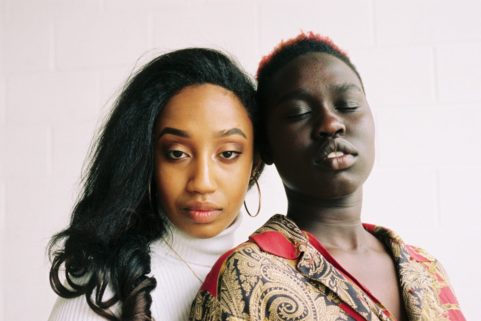 a dark-skinned short-haired woman with her eyes closed leans against a long-haired brown-skinned woman with her eyes open