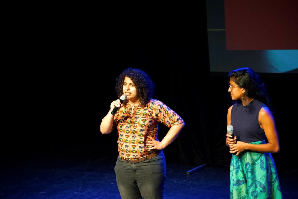 Photo of young woman on a stage speaking into a microphone, standing with a hand on her hip. Another woman stands to her right, face turned and looking at the speaker