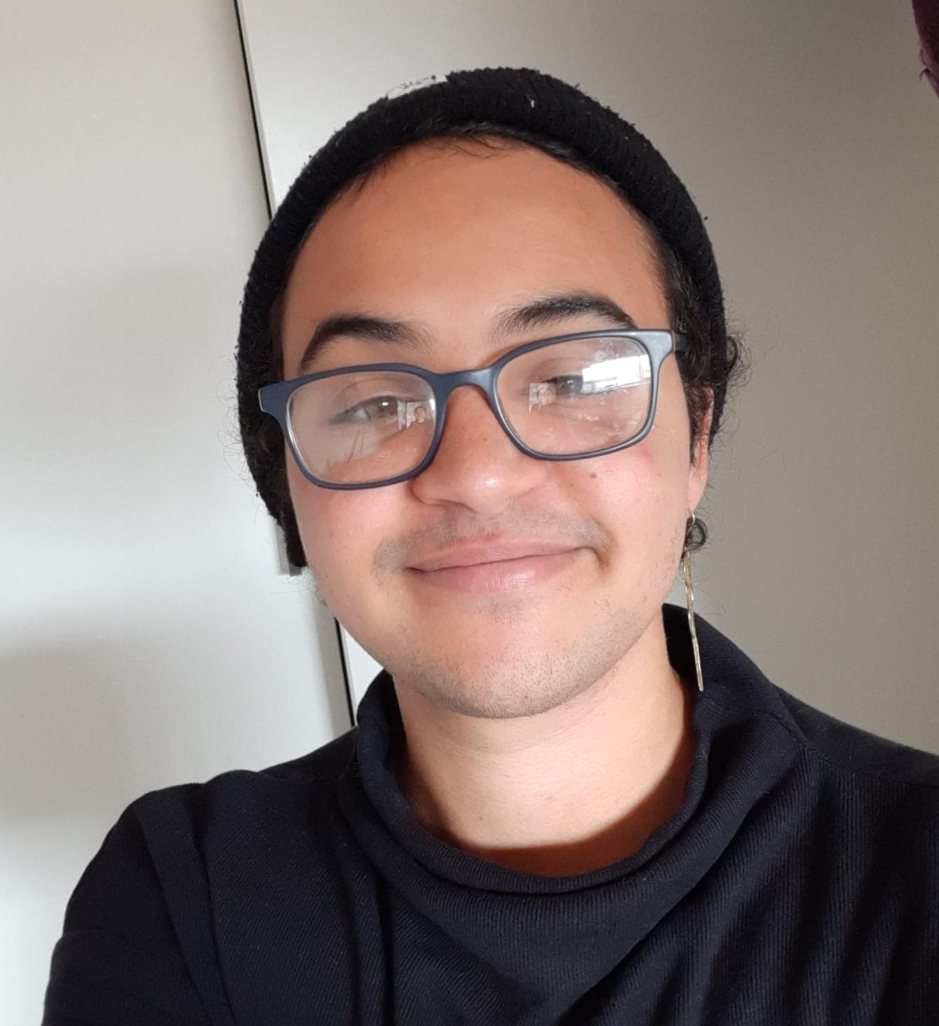 Image of a young, mixed race Afro-Anglo young man. He's wearing a black beanie, black top, and glasses. He's smiling and looking directly at the camera.